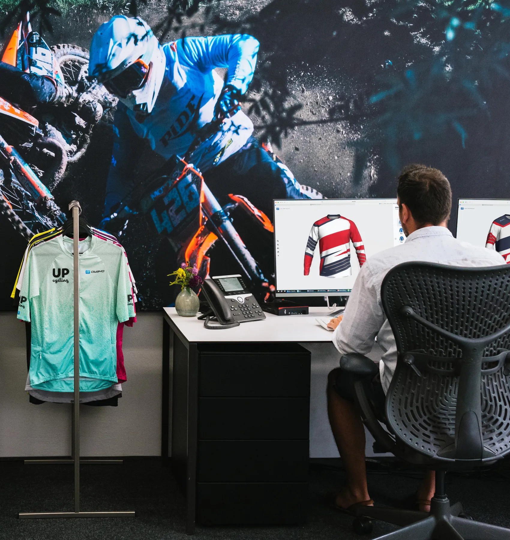 A man sits at a desk designing a customized sports jersey on a computer, with a large image of a motorcyclist and a clothing rack with various sportswear in the background.