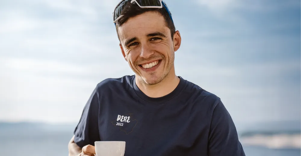 A sporty man in a navy blue T-shirt with the print 'PERE 2022' is smiling while holding a cup, set against a blurred coastal background.