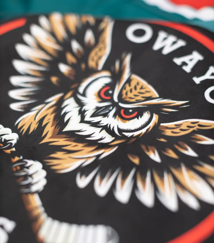 A close-up of a black sports jersey featuring a stylized owl logo holding a hockey stick, with the text 'owayo' in white lettering.