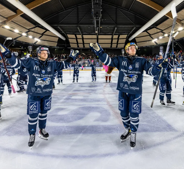 Hockey players in blue jerseys with sponsor logos celebrate on the ice, with spectators visible in the background.