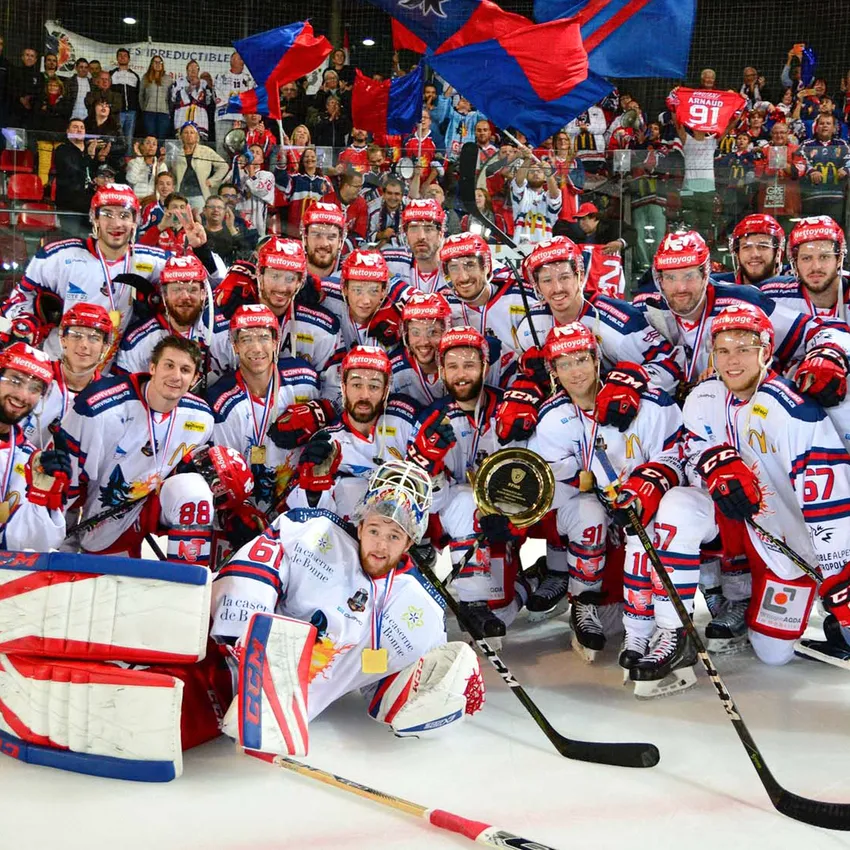 A hockey team proudly poses on the ice with medals and a trophy while fans cheer in the background.