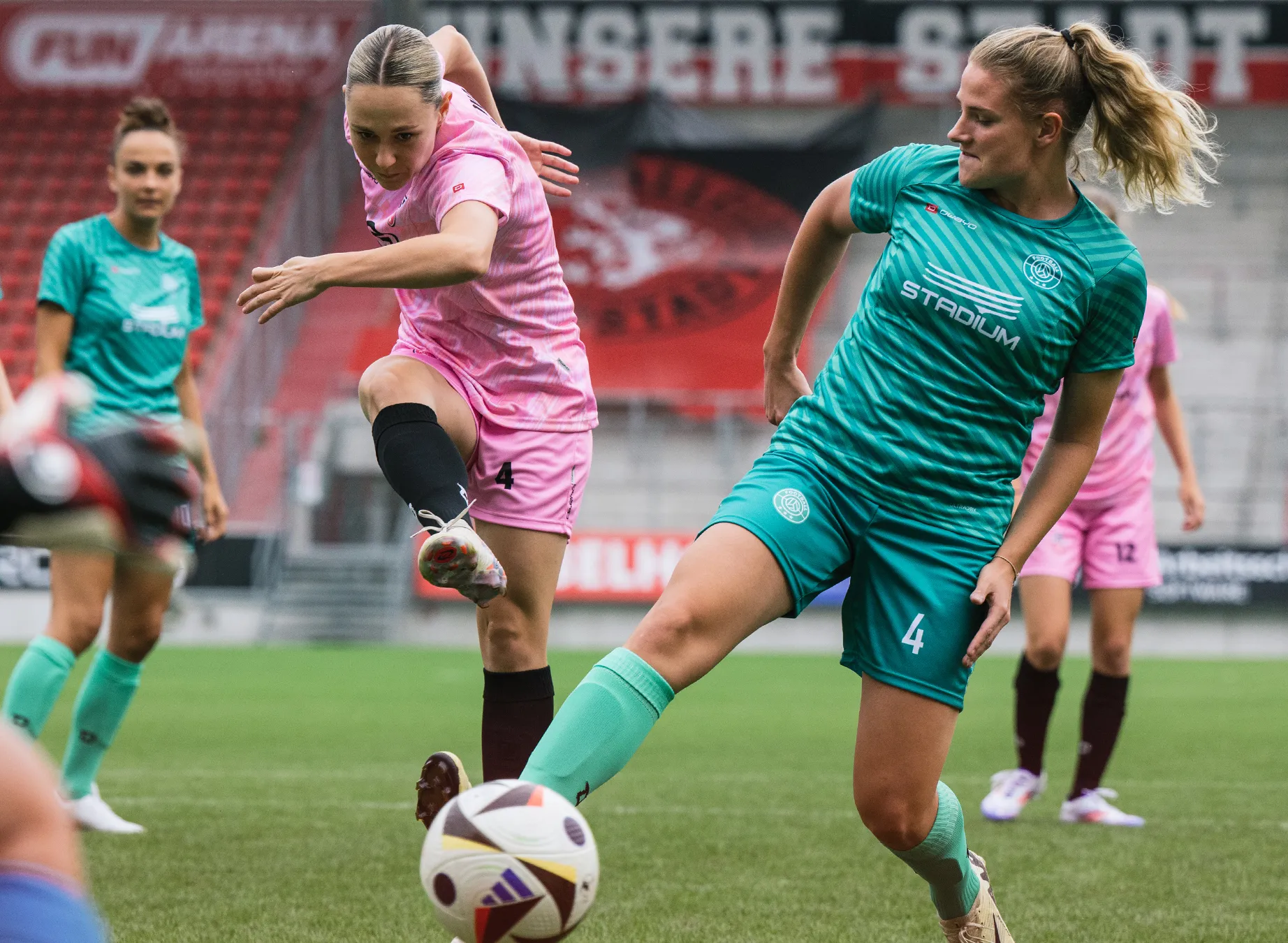 Two female soccer players in vibrant jerseys, one in pink and the other in green, are playing on a grass field.
