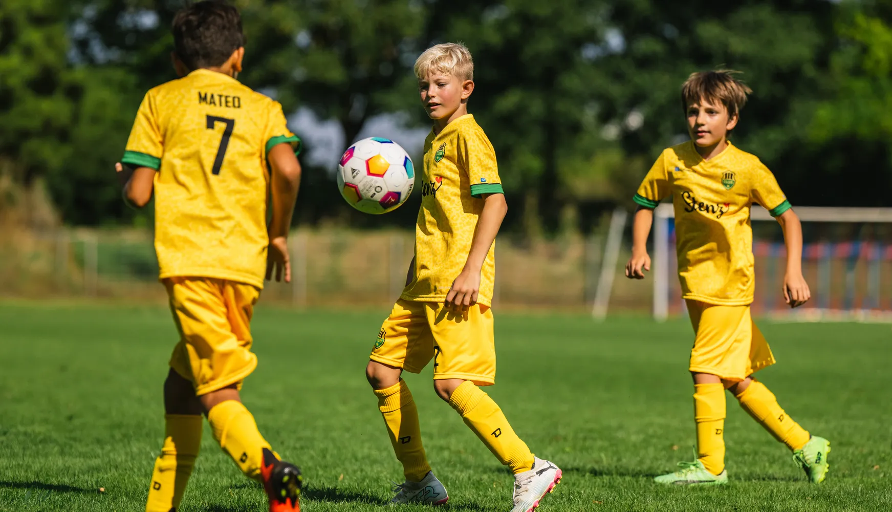 Three children in yellow soccer jerseys are playing on a grassy field with a colorful soccer ball.