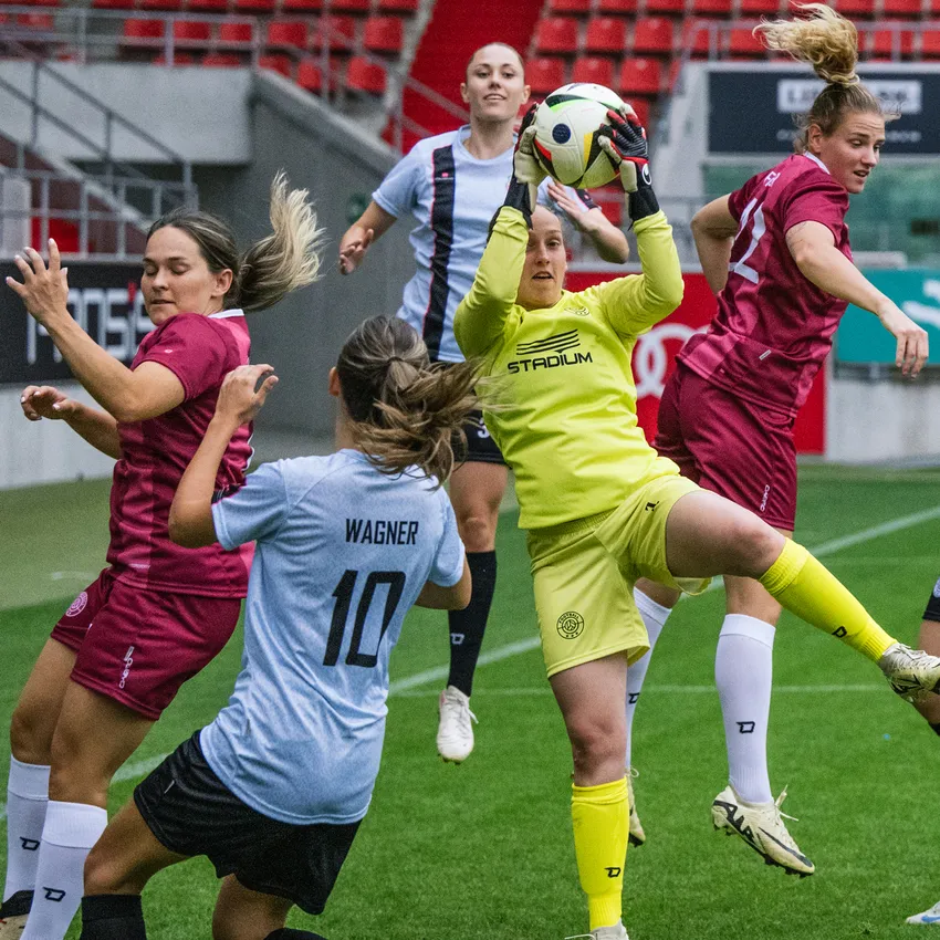 A goalkeeper in yellow sportswear catches the ball as players in different jerseys engage on the soccer field.
