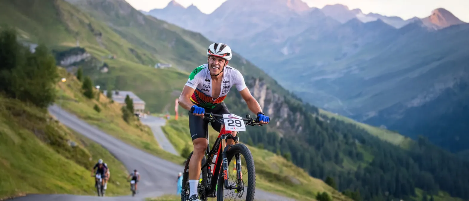 A cyclist in a white, green, and orange jersey with the number 29 rides on a mountain road in a mountainous landscape.
