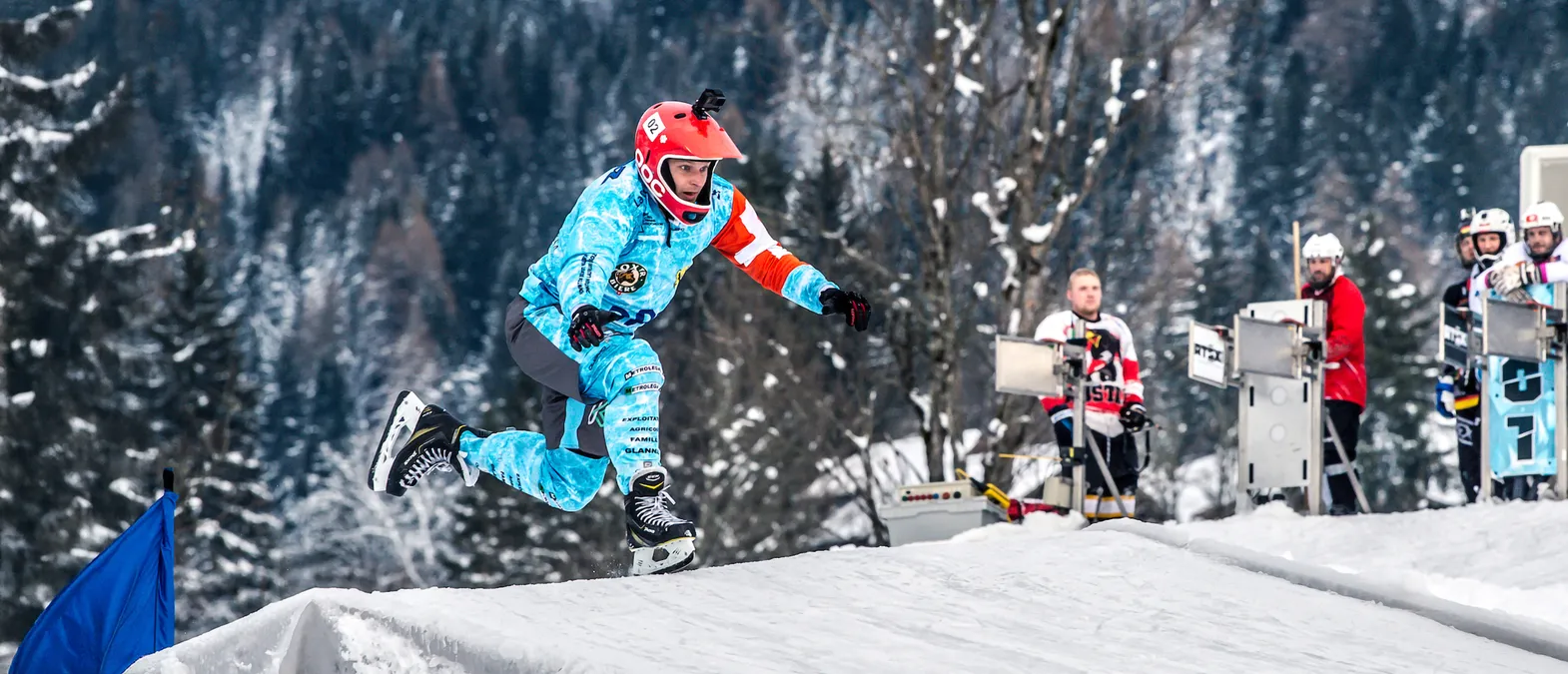 A hockey player in a blue and white custom jersey with sponsor logos and a red helmet jumps over a snow ramp on an ice rink, with other players and trees in the background.