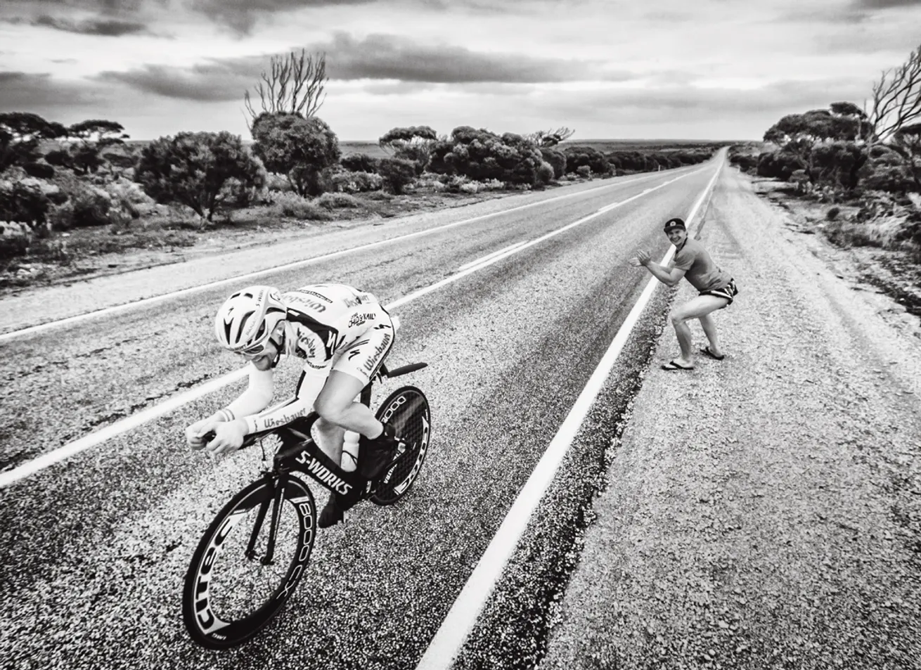 A cyclist in an aerodynamic position on a road, while a person on the roadside waves.