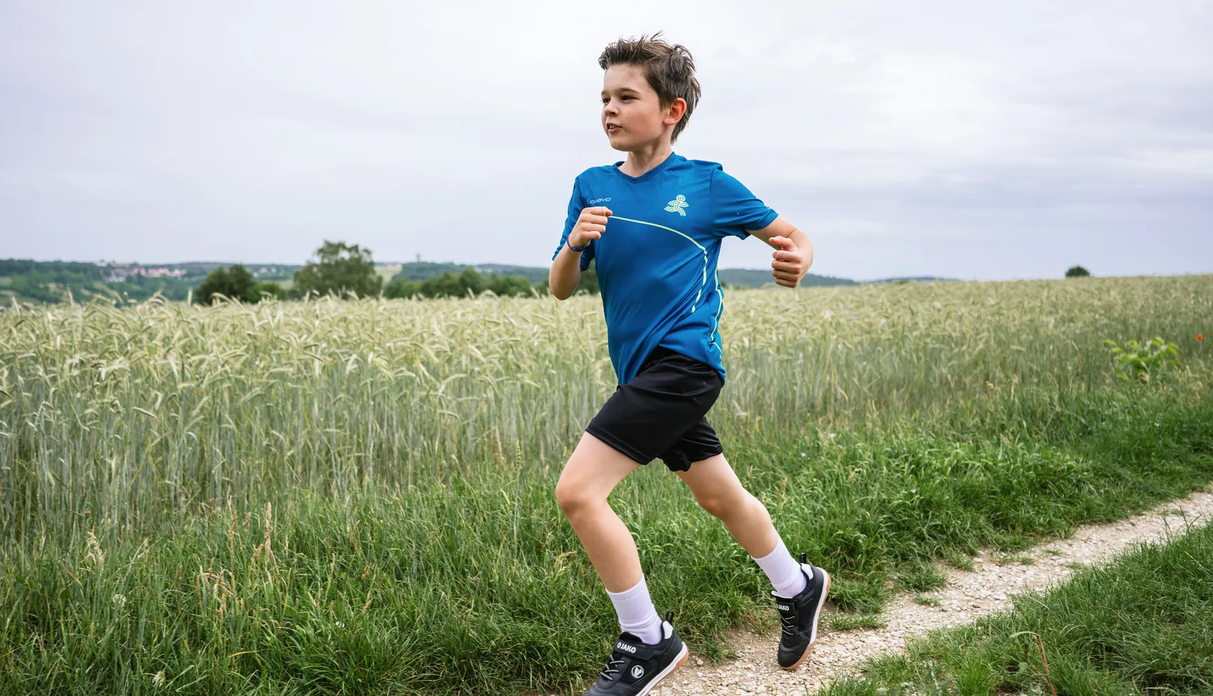 A boy runs along a path through a field, wearing a blue sports shirt and black shorts.