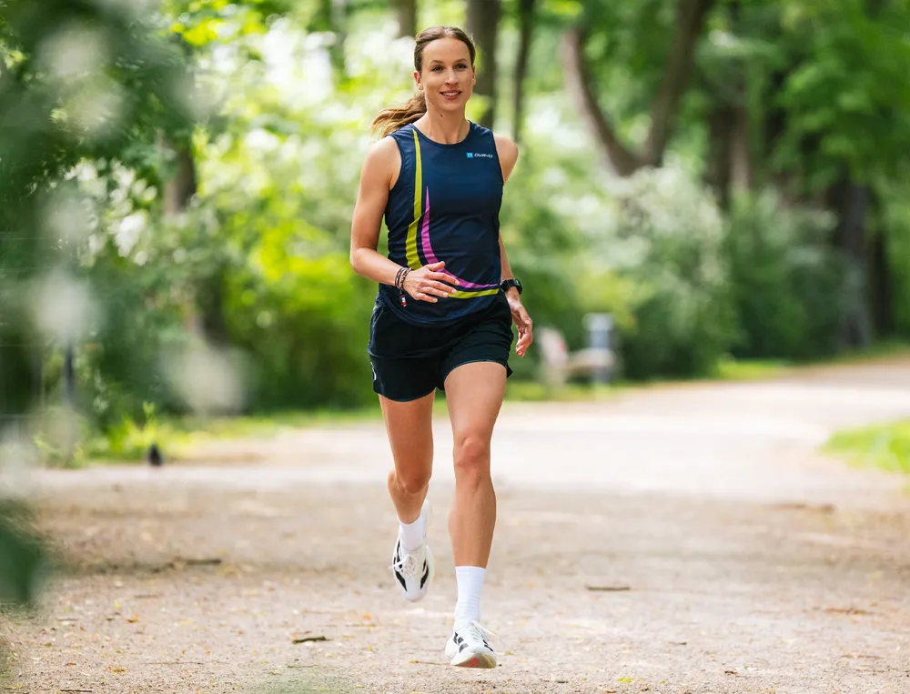 A sporty woman runs along a narrow path through a green park, wearing a blue tank top with colorful accents and shorts.