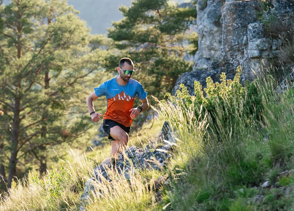 A runner in a colorful shirt and shorts sprinting over a rocky path in a green, wooded landscape.