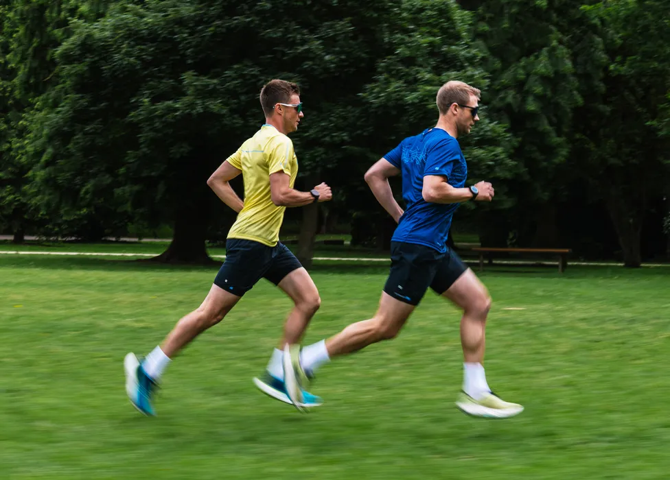Two men in sportswear are running on a grassy field, one wearing a yellow shirt and the other a blue shirt.