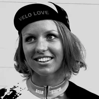 A woman with short blonde hair wears a black cycling cap with the text 'VELO LOVE' and a customized cycling jersey with the name 'Anja Sturm'.