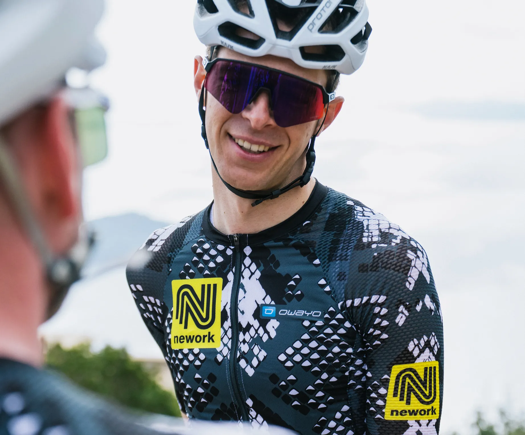 A cyclist wears a custom black and white patterned jersey with a yellow 'nework' logo and an 'owayo' logo on the chest.