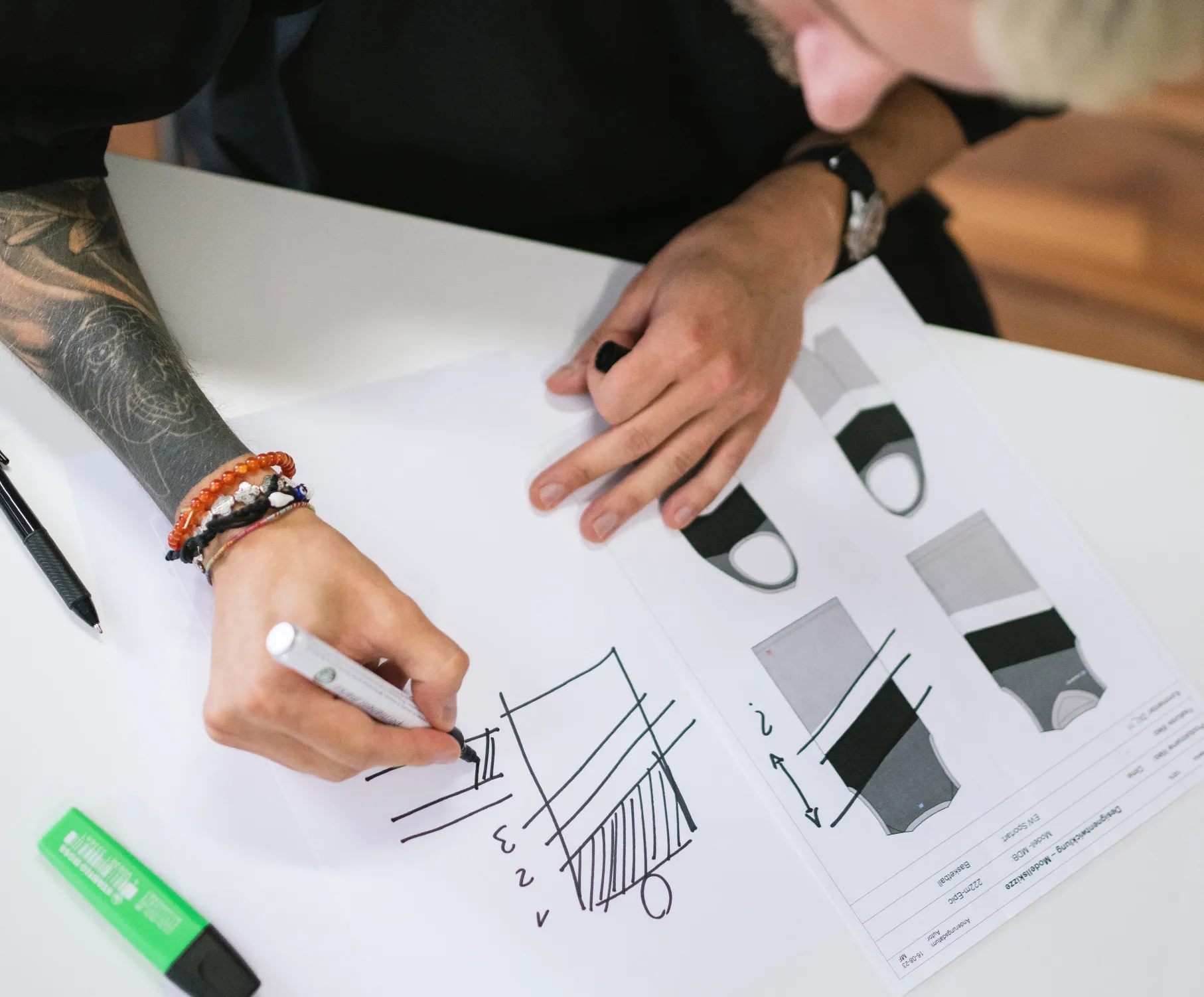 A person with a tattoo on their arm draws a design on paper with a marker, while technical drawings of sportswear lie on the table.