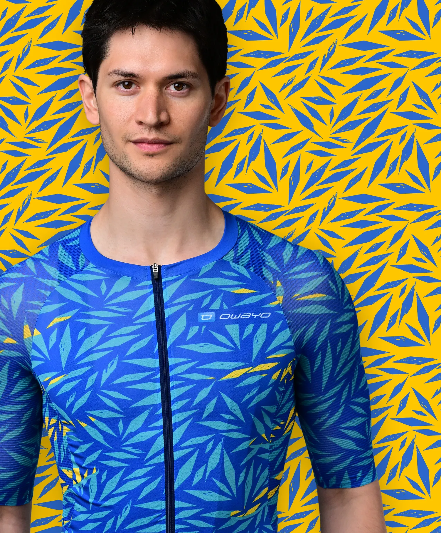 A man wears a blue, short-sleeved sport jersey with a blue and yellow leaf pattern resembling a tropical plant, against a yellow background with blue leaf patterns.