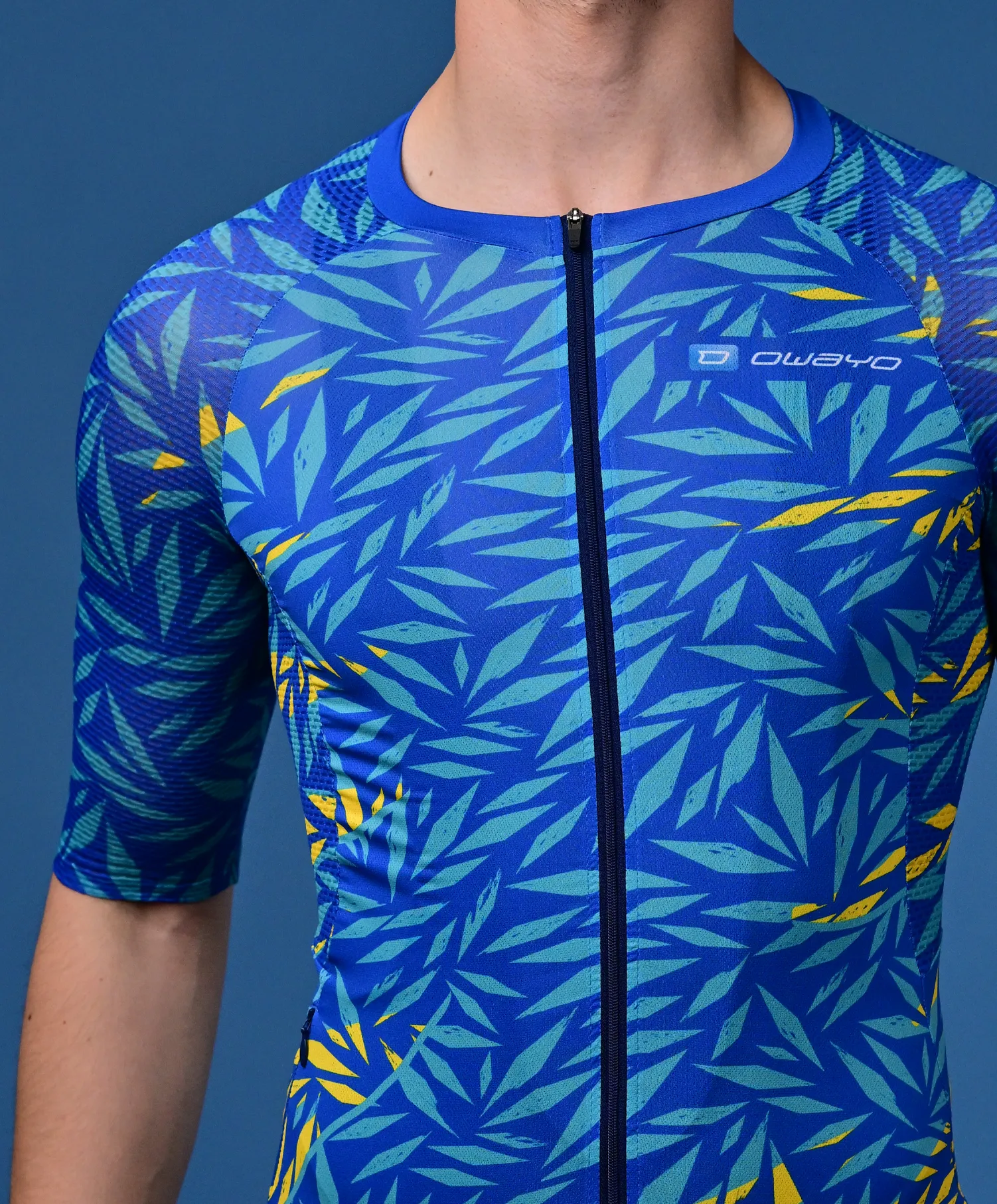 A man wears a blue, short-sleeved sports jersey with a tropical leaf pattern in teal and yellow and a front zipper.