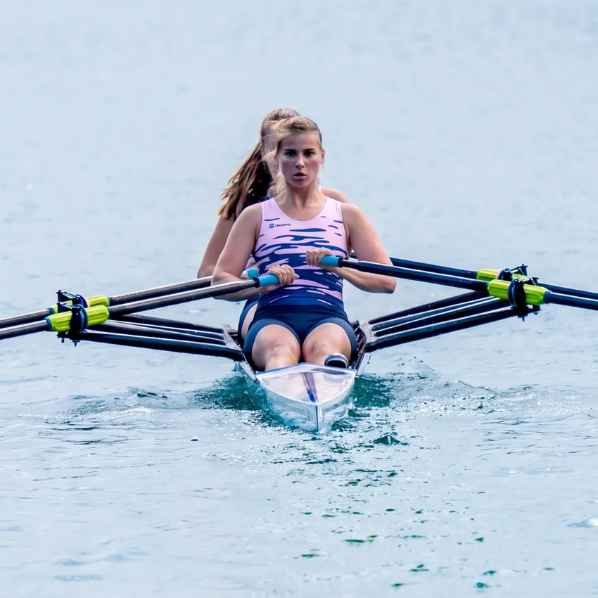Two rowers in a boat rowing on calm water, with the front rower wearing a pink sports outfit.