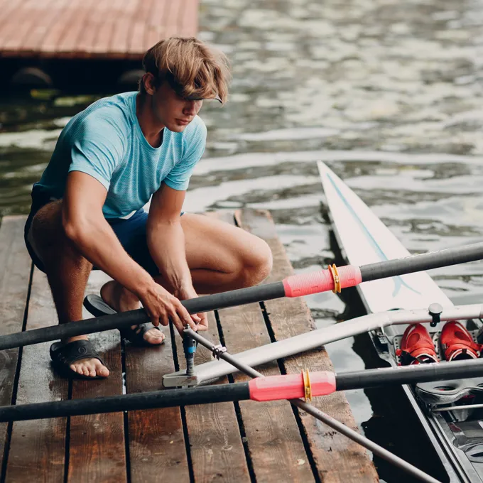 A young man in a blue sports shirt is repairing oars on a dock by the water.