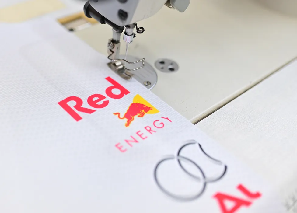 A sewing machine is stitching a white sportswear garment with a red Red Bull Energy logo and another logo.