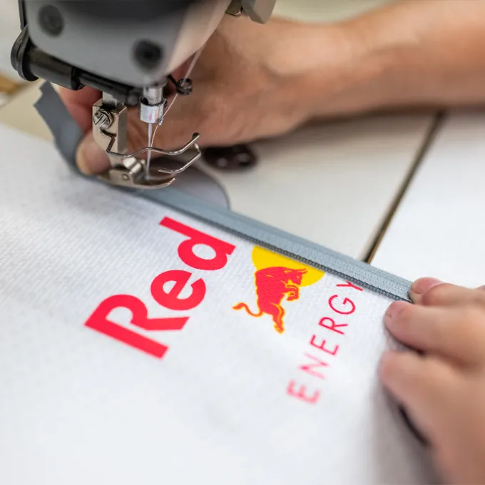 A close-up of a sewing machine stitching a white sports garment with the Red Bull logo, while a person holds the fabric.