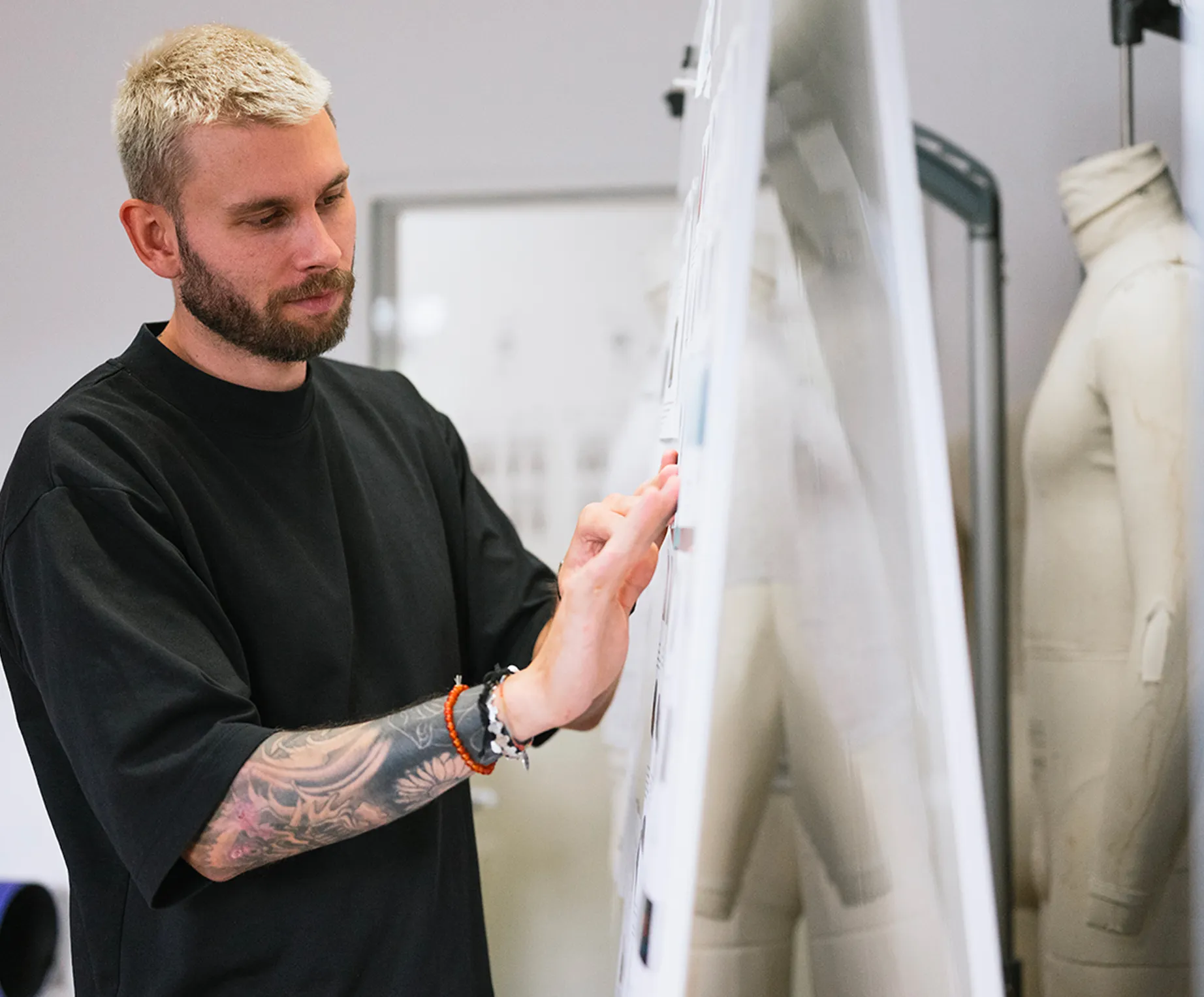 A man with blond hair and a tattoo on his arm is focused on reviewing designs on a pinboard in a design studio, with a mannequin visible in the background.